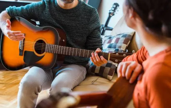 Dos personas practicando con guitarras acústicas durante una clase musical en casa, una enseñando y otra aprendiendo con atención.