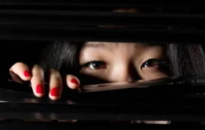 Mujer espiando a través de una persiana en un ambiente oscuro, representando la sospecha de que tu celular tiene micrófono espía.